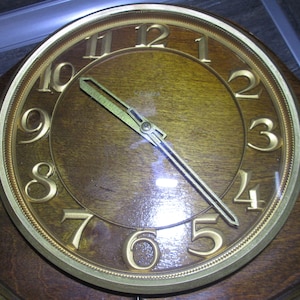 May include: A vintage wooden clock face with gold Roman numeral hour markers and gold hands. The clock face is surrounded by a gold rim with a decorative pattern. The clock face is set to 1:55.