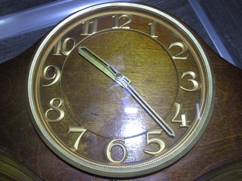 May include: A vintage wooden clock face with gold Roman numeral hour markers and gold hands. The clock face is surrounded by a gold rim with a decorative pattern. The clock face is set to 1:55.