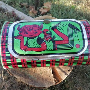 May include: Vintage metal lunchbox with a green, red, and black plaid pattern. The lid features a cartoon illustration of a girl with red hair and a monkey. The lunchbox has a metal handle and a clasp closure.