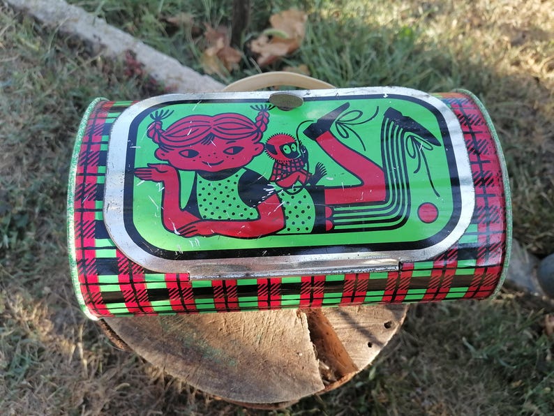 May include: Vintage metal lunchbox with a green, red, and black plaid pattern. The lid features a cartoon illustration of a girl with red hair and a monkey. The lunchbox has a metal handle and a clasp closure.