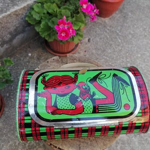 May include: Vintage metal lunchbox with a cartoon illustration of a girl and a monkey on a green background. The lunchbox has a red and black plaid pattern on the sides and a silver trim.