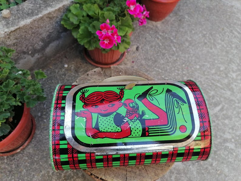 May include: Vintage metal lunchbox with a cartoon illustration of a girl and a monkey on a green background. The lunchbox has a red and black plaid pattern on the sides and a silver trim.