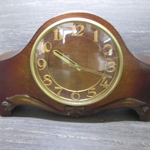 May include: A wooden mantel clock with a gold-coloured face and black hands. The clock has a clear glass cover over the face.