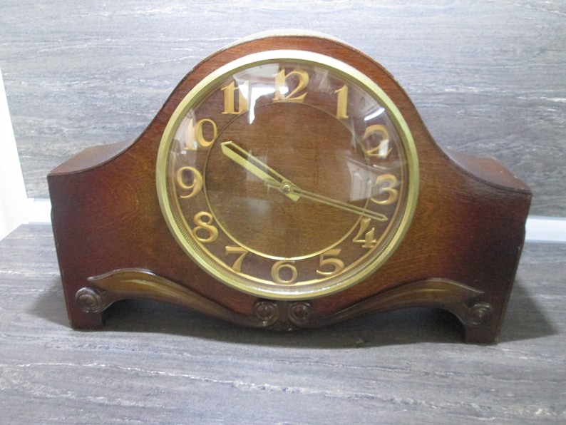 May include: A wooden mantel clock with a gold-coloured face and black hands. The clock has a clear glass cover over the face.