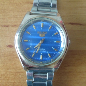 May include: A silver Seiko wristwatch with a blue face and gold-coloured hands and hour markers. The watch face also has the text "SEIKO" and "Crystal Jewels".