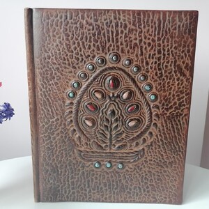 May include: A brown leather-bound book with an embossed design on the cover. The design features a central floral motif with red, tan, and turquoise accents. The book is standing upright on a white surface.