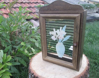 May include: A wooden cabinet with a glass door featuring a painted image of pink flowers in a blue vase. The cabinet is sitting on a tree stump.