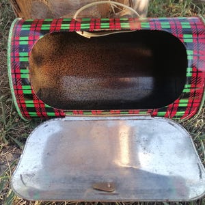 May include: Vintage metal lunchbox with a red, black, and green plaid pattern. The lunchbox is open, revealing the interior. The lid is silver-colored and has a clasp. A white handle is attached to the top.