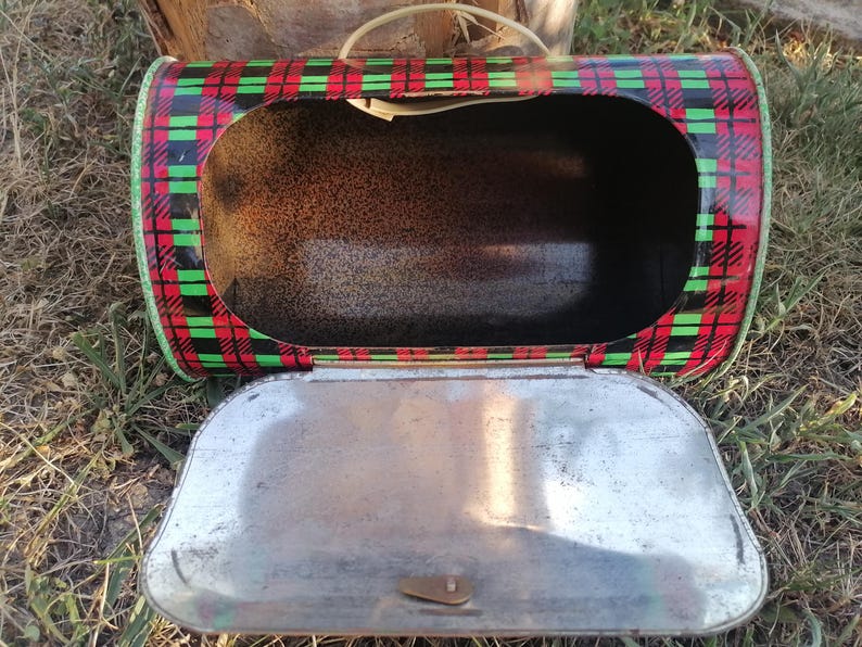 May include: Vintage metal lunchbox with a red, black, and green plaid pattern. The lunchbox is open, revealing the interior. The lid is silver-colored and has a clasp. A white handle is attached to the top.