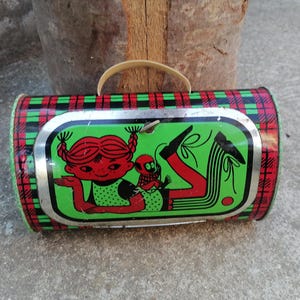 May include: Vintage cylindrical tin lunchbox with a plaid pattern in red, green, and black. The front features a cartoon illustration of a girl with red hair and a small companion on a green background.