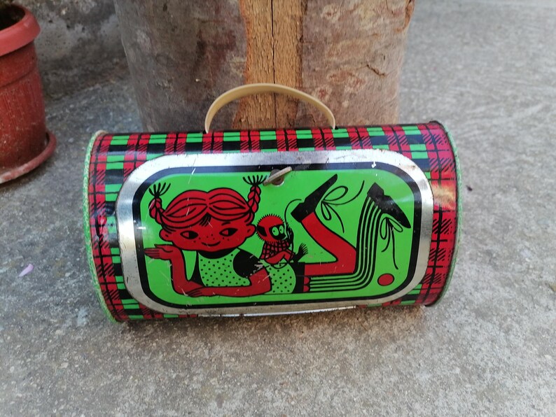 May include: Vintage cylindrical tin lunchbox with a plaid pattern in red, green, and black. The front features a cartoon illustration of a girl with red hair and a small companion on a green background.