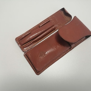 Könnte beinhalten: Zwei braune Lederetuis für Stifte oder Bleistifte. Das Etui links hat drei Fächer für Schreibgeräte. Das Etui rechts hat eine Klappe, die sich über die Oberseite schließt.