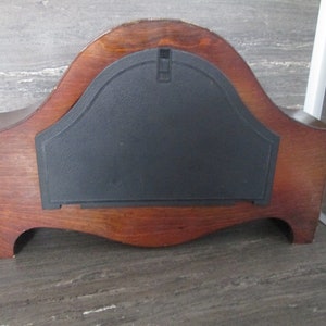 May include: The back of a wooden clock with a black plastic cover. The clock has a curved shape and a dark wood finish.