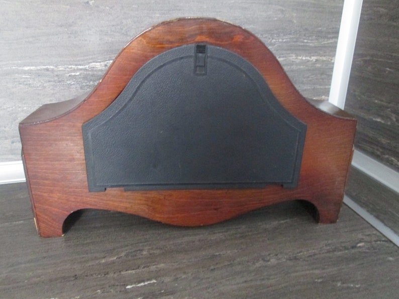 May include: The back of a wooden clock with a black plastic cover. The clock has a curved shape and a dark wood finish.