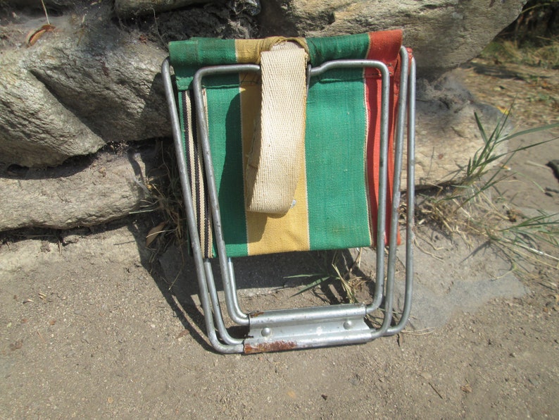 K&ouml;nnte beinhalten: Ein Klappstuhl mit einer Sitzfl&auml;che und R&uuml;ckenlehne aus gr&uuml;nem, gelbem und rotem gestreiftem Stoff. Der Stuhl ist aus Metall und hat einen wei&szlig;en Trageriemen.