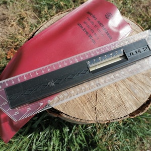 Puede incluir: Una regla de plástico transparente con una regla negra unida a ella. La regla tiene marcas en centímetros y pulgadas. La regla está etiquetada como "JL2" y "ЛИНЕЙКА ИНЕРЦИОННАЯ".