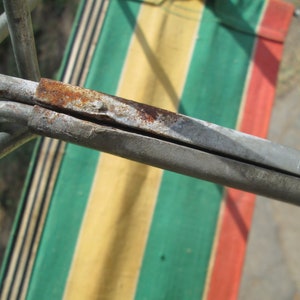 K&ouml;nnte beinhalten: Nahaufnahme eines Metallrahmens mit sichtbarem Rost, der an einem gestreiften Stoff mit gr&uuml;nen, gelben und roten Streifen befestigt ist.