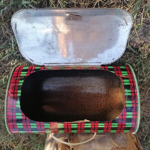 May include: Vintage metal bread box with an open lid. The box has a red, black, and green plaid pattern on the exterior. The interior is dark, and the lid is silver. A handle is visible on the bottom.