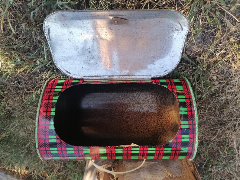 May include: Vintage metal bread box with an open lid. The box has a red, black, and green plaid pattern on the exterior. The interior is dark, and the lid is silver. A handle is visible on the bottom.