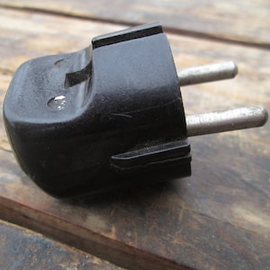 Puede incluir: Un enchufe eléctrico negro de dos clavijas con un cuerpo redondeado y texturado y clavijas de metal plateado.