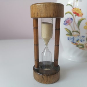 Könnte beinhalten: Eine Sanduhr aus Holz mit einer klaren Glasbirne, die mit Sand gefüllt ist. Die Sanduhr ist etwa 15 cm hoch und hat eine natürliche Holzoptik.