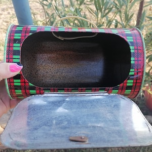 May include: A vintage metal lunchbox with a red, black, and green plaid pattern. The lunchbox has a hinged lid and a small handle. The interior is dark, and the lid is open, revealing the inside.
