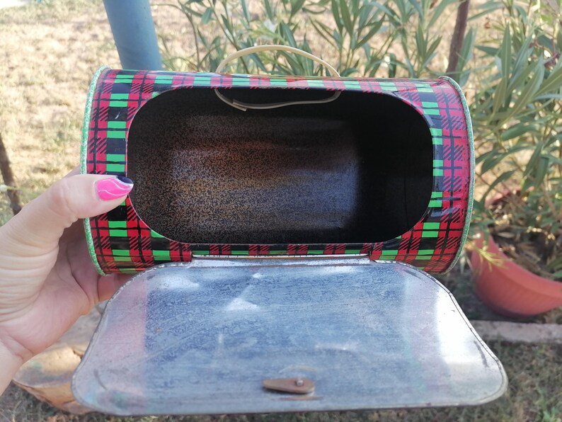 May include: A vintage metal lunchbox with a red, black, and green plaid pattern. The lunchbox has a hinged lid and a small handle. The interior is dark, and the lid is open, revealing the inside.