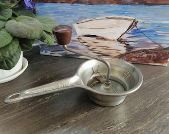 May include: A silver food mill with a brown plastic handle. The mill has a strainer and a crank. The mill is on a wooden surface.