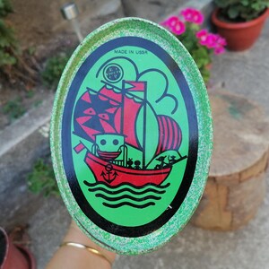 May include: Oval-shaped vintage tin with a green border and a central illustration of a red sailboat with black sails and a black hull. The tin has the text "MADE IN USSR" and a logo.