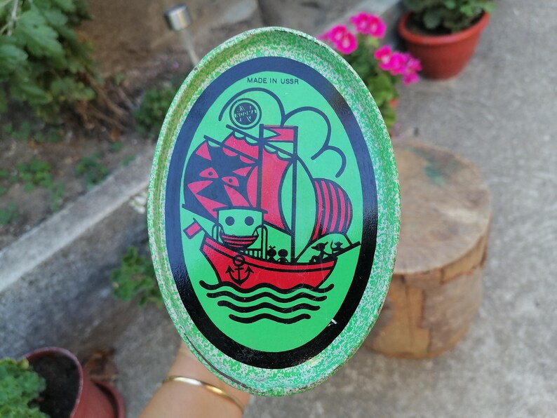 May include: Oval-shaped vintage tin with a green border and a central illustration of a red sailboat with black sails and a black hull. The tin has the text "MADE IN USSR" and a logo.