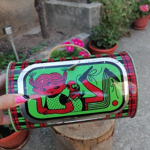 May include: Vintage metal lunchbox with a red, green, and black plaid pattern. The lid features a cartoon illustration of a girl and a creature in red, black, and green. The lunchbox has a metal handle.