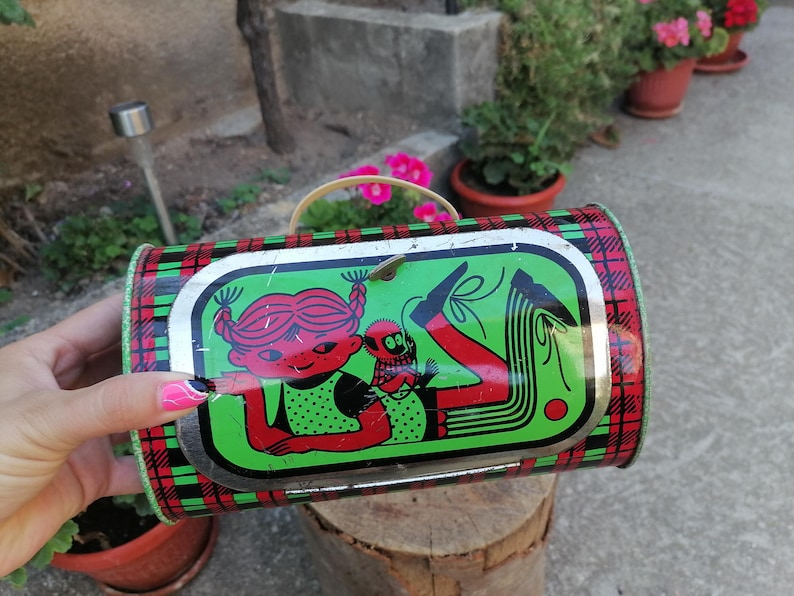 May include: Vintage metal lunchbox with a red, green, and black plaid pattern. The lid features a cartoon illustration of a girl and a creature in red, black, and green. The lunchbox has a metal handle.