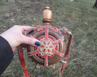 Könnte beinhalten: Eine Holzflasche mit einem Lederriemen und roten und weißen Zierstichen. Die Flasche hat einen Holzverschluss.