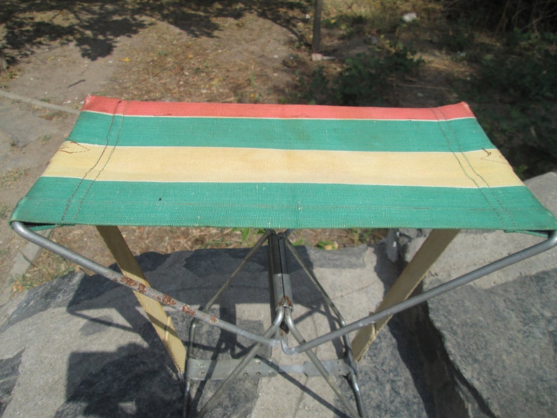 K&ouml;nnte beinhalten: Ein klappbarer Campinghocker mit einer Sitzfl&auml;che aus gr&uuml;nem, gelbem und rotem gestreiftem Stoff. Der Hocker ist aus Metall und hat ein schlichtes, funktionales Design.