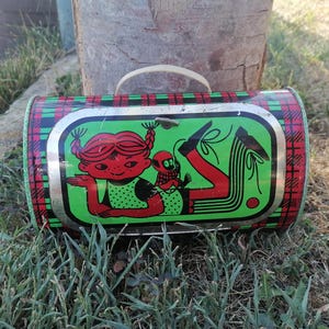 May include: Vintage metal lunchbox with a red, green, and black plaid pattern. The front features a cartoon illustration of a girl with red hair and a small creature on a green background. The lunchbox has a white handle.