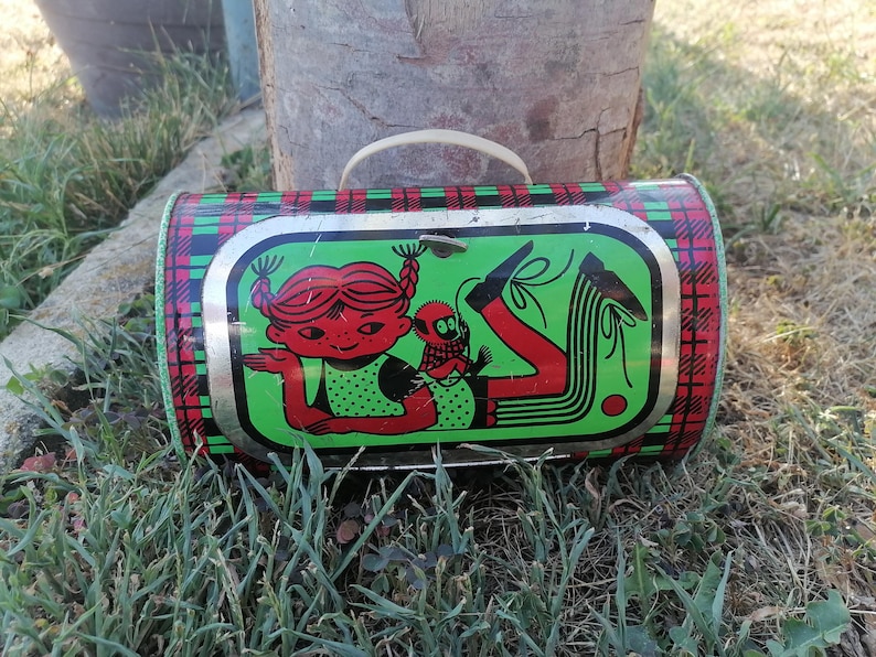 May include: Vintage metal lunchbox with a red, green, and black plaid pattern. The front features a cartoon illustration of a girl with red hair and a small creature on a green background. The lunchbox has a white handle.