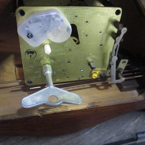 May include: Close-up of a clock mechanism with a winding key. The mechanism is made of brass and has a clear plastic cover. The clock is labelled "VISIA" and "OWN 1".