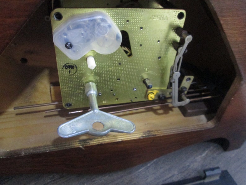 May include: Close-up of a clock mechanism with a winding key. The mechanism is made of brass and has a clear plastic cover. The clock is labelled "VISIA" and "OWN 1".