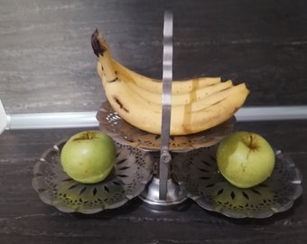 May include: A silver two-tiered fruit stand with a handle. The top tier holds a bunch of yellow bananas. The bottom tier holds two green apples.