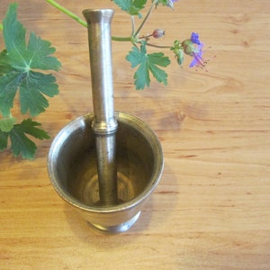 May include: A silver mortar and pestle set. The mortar is a bowl with a rounded bottom and a wide opening. The pestle is a long, cylindrical rod with a rounded end. The mortar and pestle are sitting on a wooden surface.