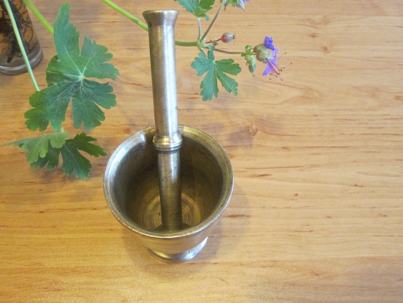 May include: A silver mortar and pestle set. The mortar is a bowl with a rounded bottom and a wide opening. The pestle is a long, cylindrical rod with a rounded end. The mortar and pestle are sitting on a wooden surface.