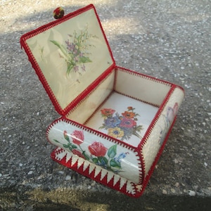 Op de afbeelding: Een vintage, witte, gehaakte doos met een rode rand en bloemmotieven. De doos is open en de binnenkant is bekleed met wit papier met een bloemmotief.