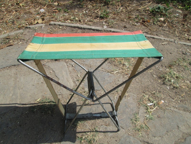 K&ouml;nnte beinhalten: Ein klappbarer Metallstuhl mit einem gestreiften Stoffbezug in Rot, Gr&uuml;n und Beige. Der Stuhl ist aus Metall und hat ein schlichtes Design.