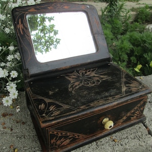 May include: An antique black wooden vanity box with a hinged mirror and a drawer. The box features decorative floral and geometric carvings with a light brown accent. The drawer has a cream-coloured knob. The mirror is rectangular.