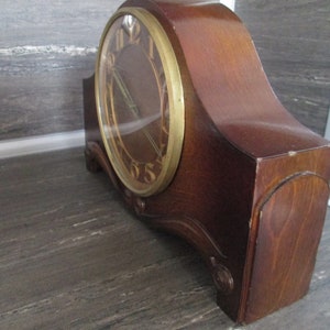May include: A vintage wooden mantel clock with a gold-toned face and Roman numeral hour markers. The clock has a curved wooden case with a dark wood finish.