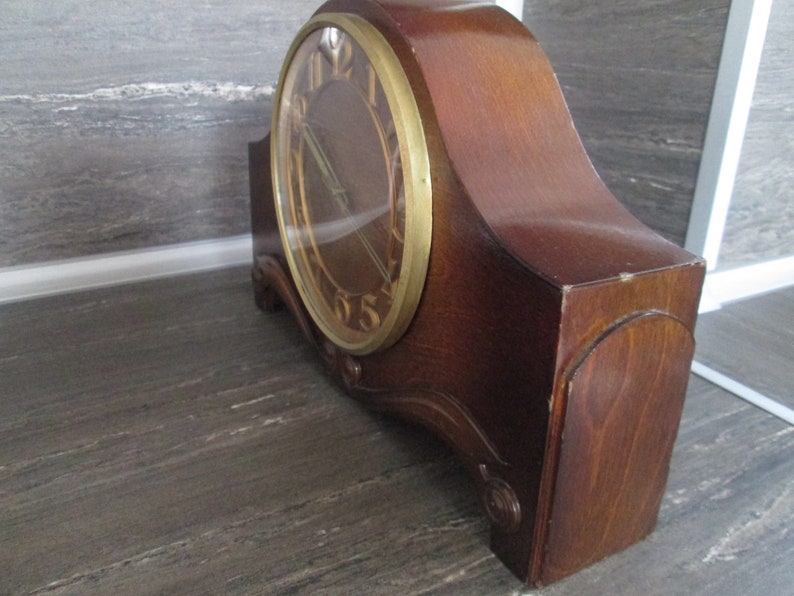 May include: A vintage wooden mantel clock with a gold-toned face and Roman numeral hour markers. The clock has a curved wooden case with a dark wood finish.