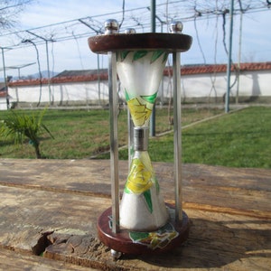 May include: A small, decorative hourglass with a wooden base and a clear glass bulb. The bulb is painted with yellow flowers and green leaves. The hourglass is sitting on a wooden table.