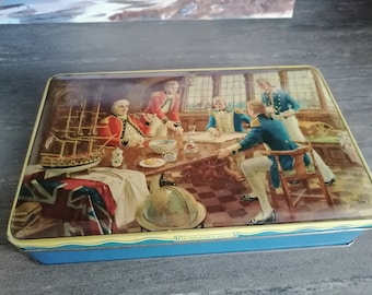 May include: A vintage tin box with a blue lid and a colourful illustration of a group of men in naval uniforms gathered around a table. The scene depicts a ship in the background and a globe on the table. The text "In Nelson's Days" is printed on the front of the box.