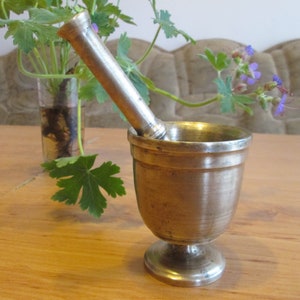 May include: A silver mortar and pestle set on a wooden table. The mortar is a bowl with a rounded bottom and a flared lip. The pestle is a long, cylindrical rod with a rounded end.