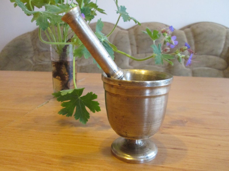 May include: A silver mortar and pestle set on a wooden table. The mortar is a bowl with a rounded bottom and a flared lip. The pestle is a long, cylindrical rod with a rounded end.